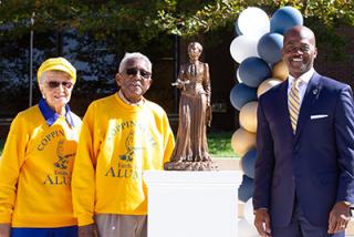 James "Winky" and Florine "Peaches" Camphor wear yellow Coppin alumni sweatshirts and sunglasses while standing next to President Anthony Jenkins and a miniature bronze statue of Fanny Jackson Coppin
